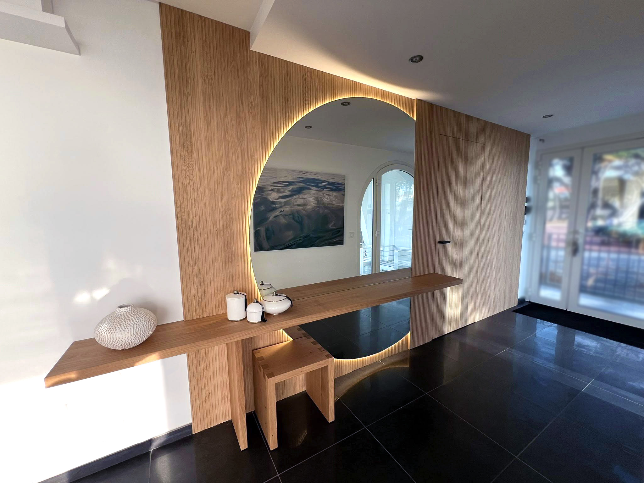 Entrée habillée de panneaux en chêne rainuré, avec console-bureau suspendue et miroir circulaire rétroéclairé.